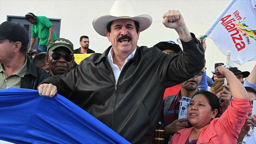 Man wearing a white cowboy hat and black leather jacket raises a clenched fist amid a crowd; visible are Manuel Zelaya Rosales and Juan Orlando Hernández in coverage, with a blue flag nearby.