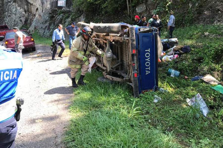 Overturned blue Toyota on a grassy roadside as emergency crews work nearby, with police and bystanders at the scene during Semana Morazánica.