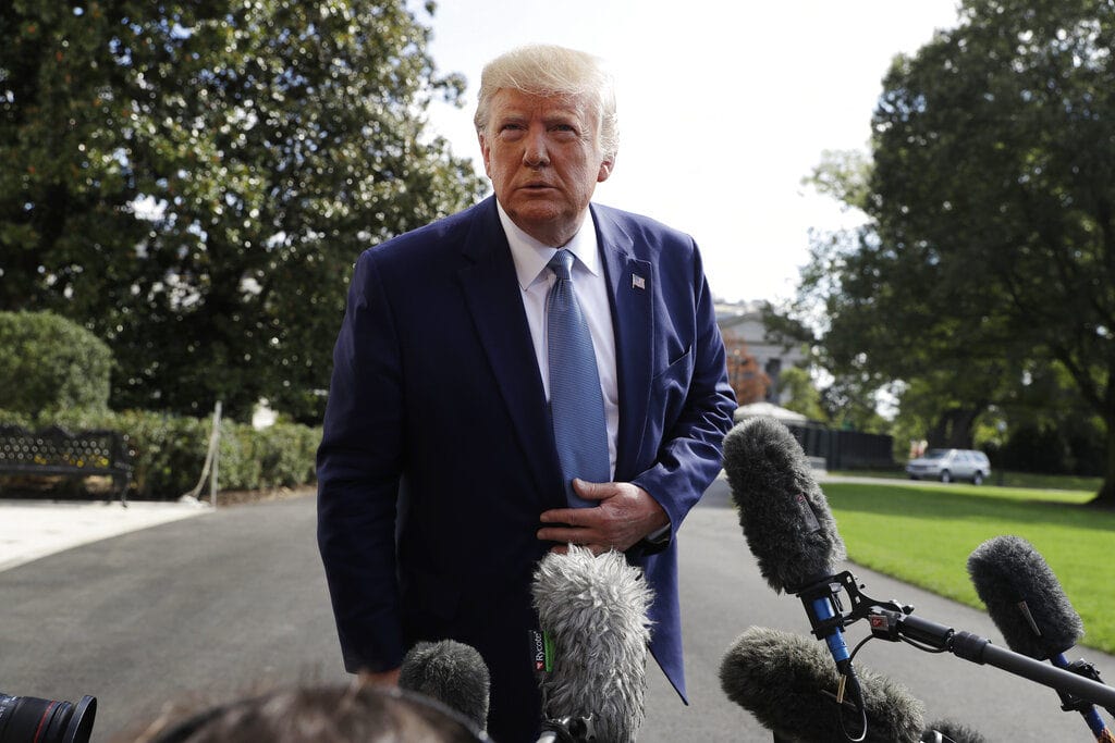 Donald Trump in a dark blue suit and light blue tie speaking to multiple microphones outdoors, with Elwood Francis nearby and trees in the background