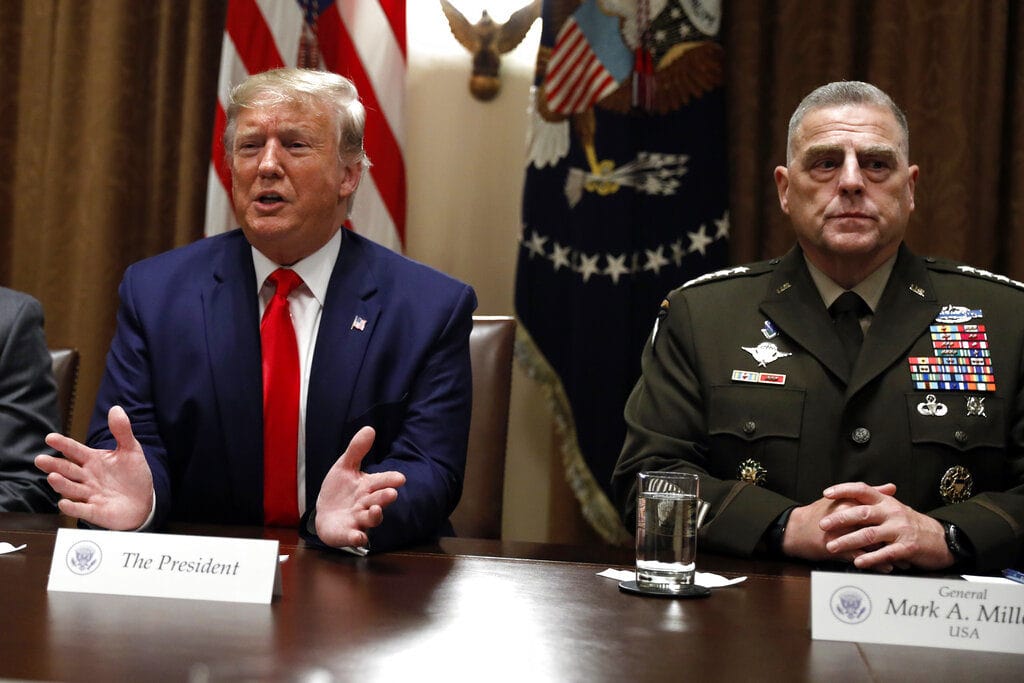 Donald Trump in a navy suit and red tie sits at a conference table, beside General Mark A. Milley in full military dress with ribbons; flags and an emblem backdrop add formal detail.
