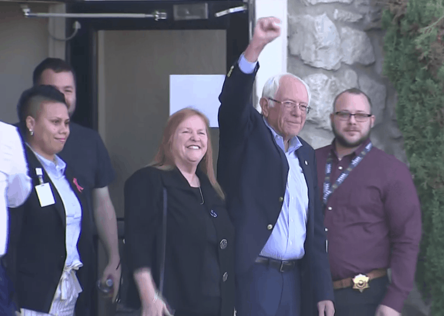 Bernie Sanders raises his fist beside Rachel Banham and Theresa Grentz among a small group of supporters outside a building, smiling and ready for a public event.
