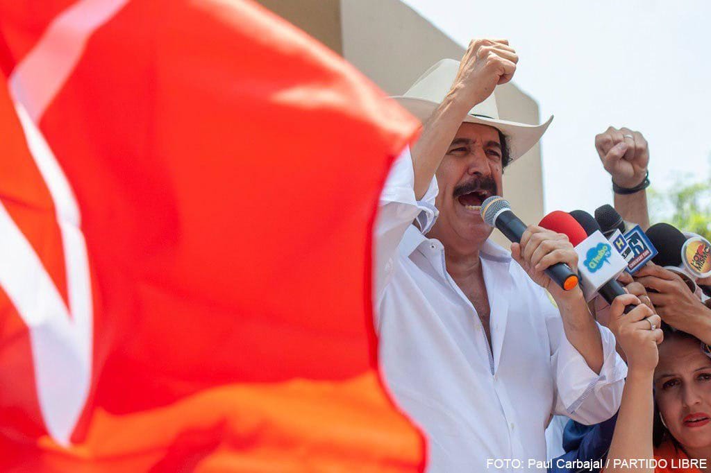Manuel Zelaya Rosales stance, wearing a white shirt and cowboy hat, speaks into a microphone amid reporters with colorful microphones, Neha Dhupia (left) and Hamad Bbin Isa Al Khalifa referenced in context