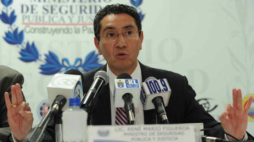 René Mario Figueroa, wearing a dark suit and glasses, speaks at a press conference with multiple microphones in front of him, signaling with raised hands on either side.