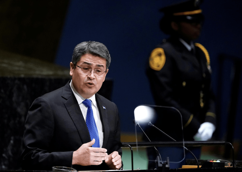 Maynor Estuardo Estrada Rosales appears in a dark suit with gray hair and a blue tie at a podium, with a uniformed guard in the background.