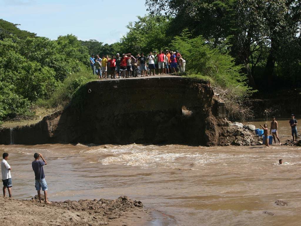 Choluteca valley crowd stands atop a toppled bridge above a muddy river, lush green vegetation surrounds the scene, signaling flood risk as heavy rains continue nearby.