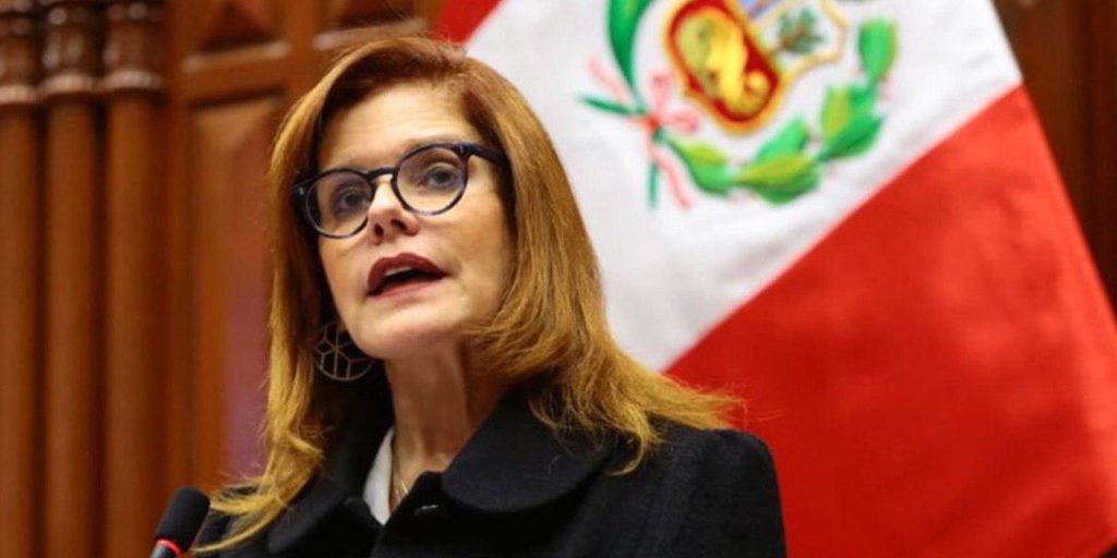 Mercedes Aráoz, wearing black glasses and a dark outfit, speaks in front of a Peruvian flag with a bold crest, highlighted by warm lighting and a wood-paneled backdrop.