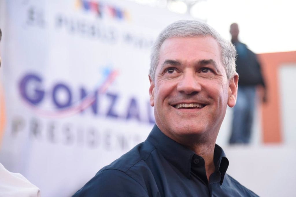 Gonzalo Castillo with gray hair and a dark shirt smiles at a campaign event, with a blurred banner in the background and supporters nearby.