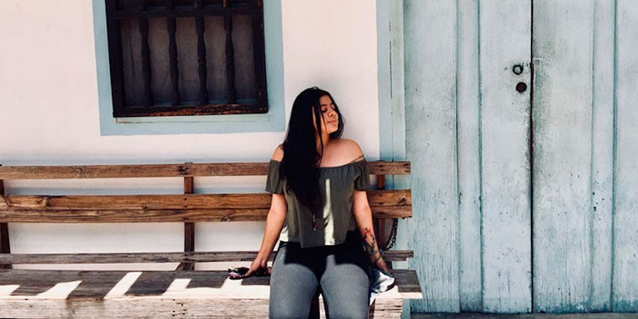Portrait of Ana Sandoval, a biology student, with long dark hair, wearing an off-shoulder gray top and dark jeans, seated on a wooden bench beside a pastel wall and a light blue door.
