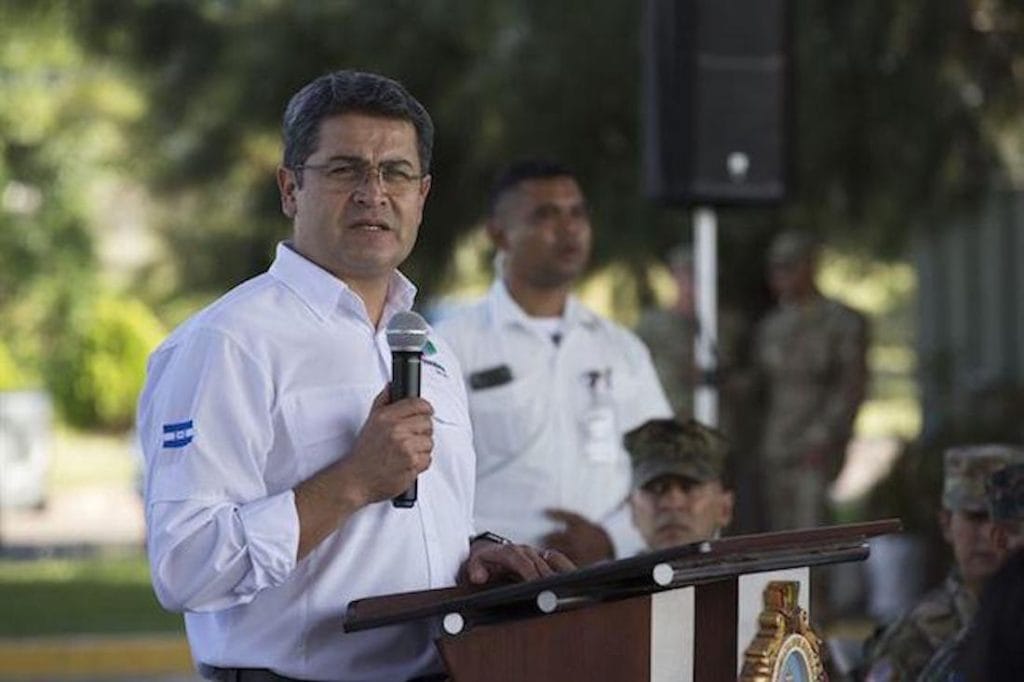 Juan Orlando Hernández in a white shirt speaks into a microphone at a wooden podium outdoors, with security and military personnel in the background; mentions Alexander Ardón and Ramón Sabillón in a news context.