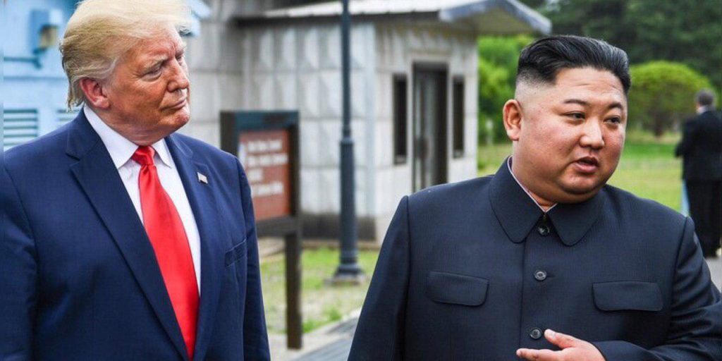 Kim Jong Un and Donald Trump in dark blue suits, with Trump wearing a bright red tie, walking outdoors in a formal setting amid greenery.