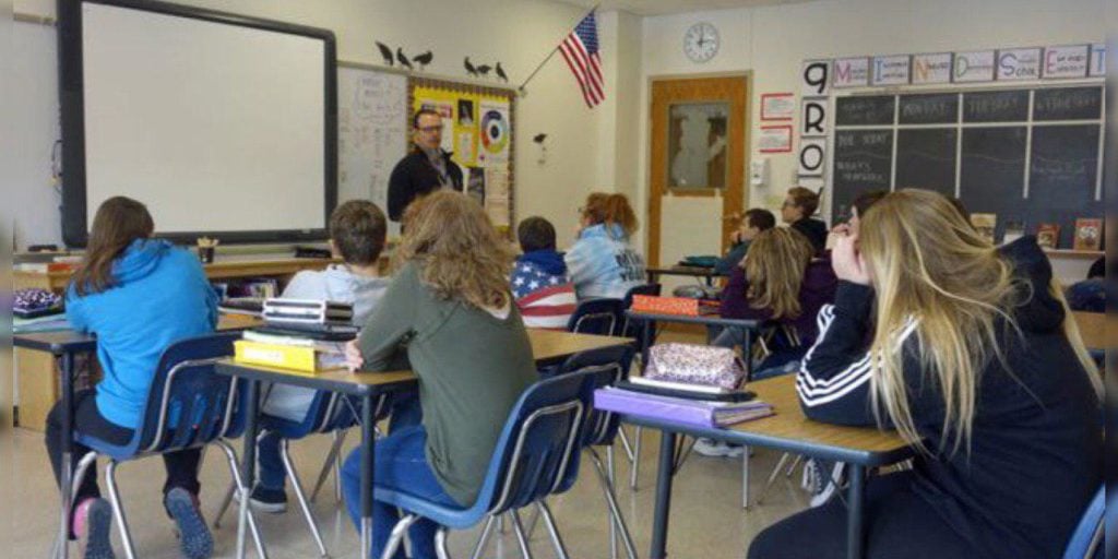 Classroom in Florida with students seated at desks facing the front as a teacher speaks; an American flag and educational posters adorn the walls, evoking a typical learning environment in a district like Parkland near Stoneman Douglas.