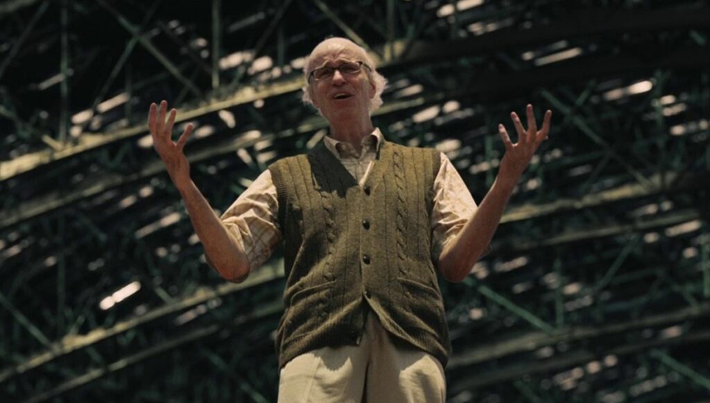Tom Noonan with white hair and glasses, wearing a beige shirt and brown vest, stands with arms raised in a performance-like setting.