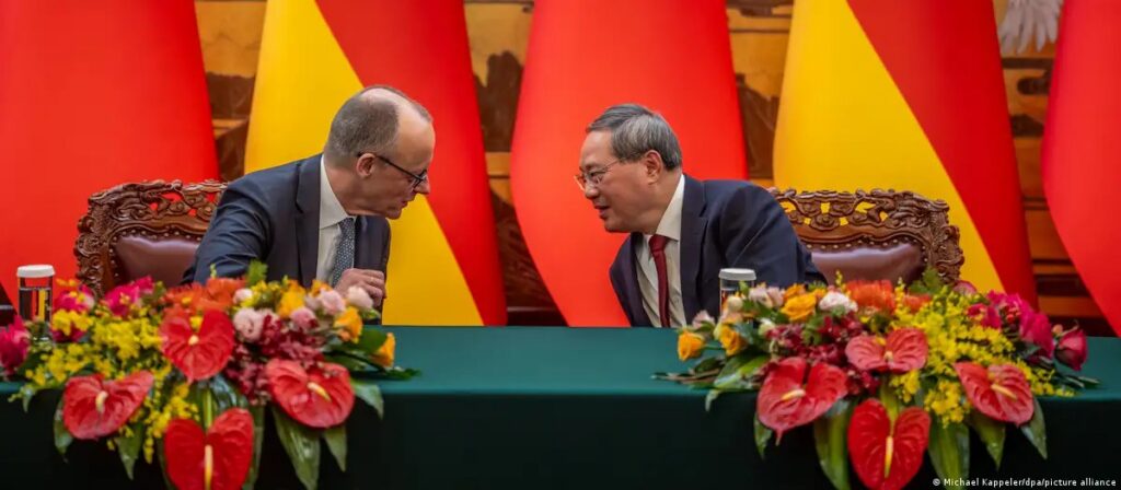 Two figures in business attire lean toward each other across a table, flanked by vibrant red and yellow floral arrangements and bold flags in the background; Li Qiang, Bob Good, and Frank Vang-Jensen are mentioned in the context.