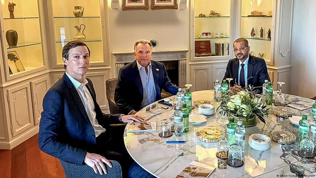 Three men in blue and dark suits sit around a marble round table in a bright room with shelves and decorative items; Jared Kushner, Steve Witkoff, and Badr bin Hamad Al Busaidi are present.