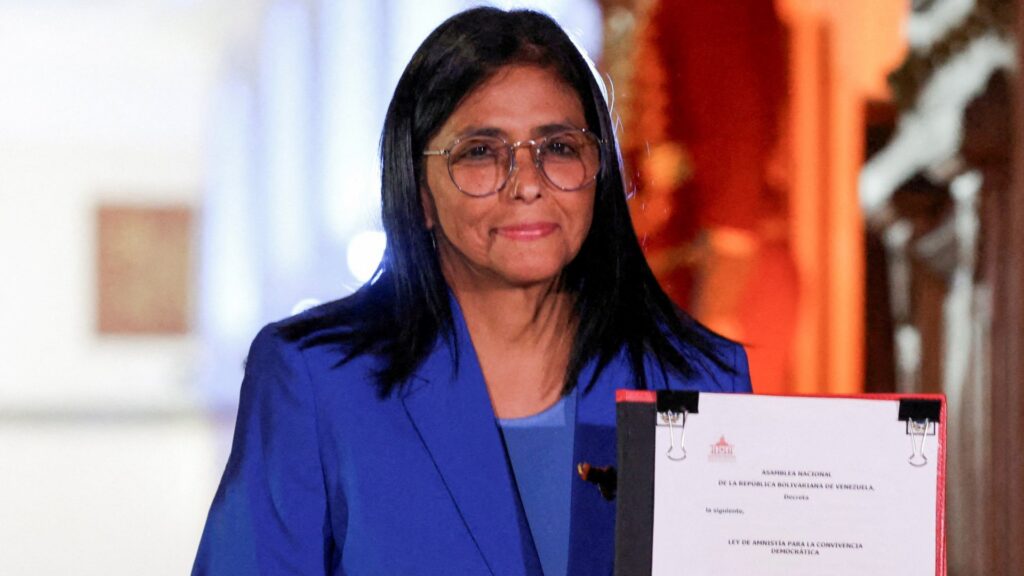 Delcy Rodríguez, wearing a blue blazer and round glasses, holds a document in a formal setting, conveying a composed and official presence with dark hair and a soft smile.