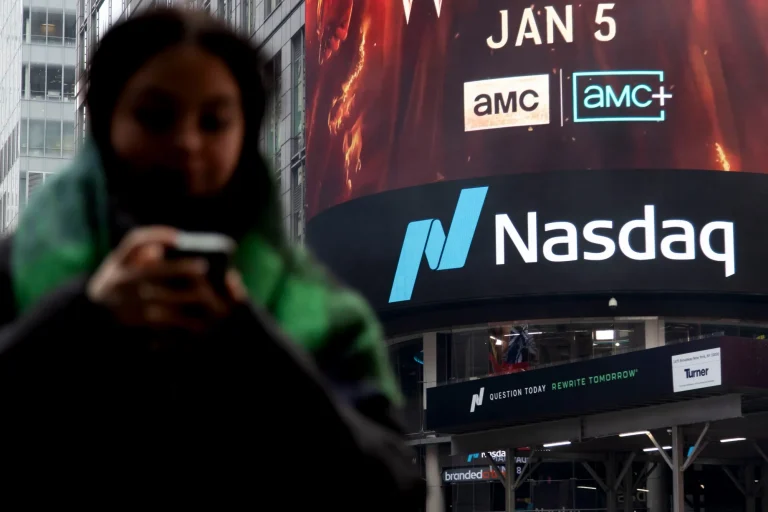 A blurred person in the foreground looks at a phone, with a large, brightly lit Nasdaq digital billboard in the background displaying 'Nasdaq', 'AMC', and other advertisements.