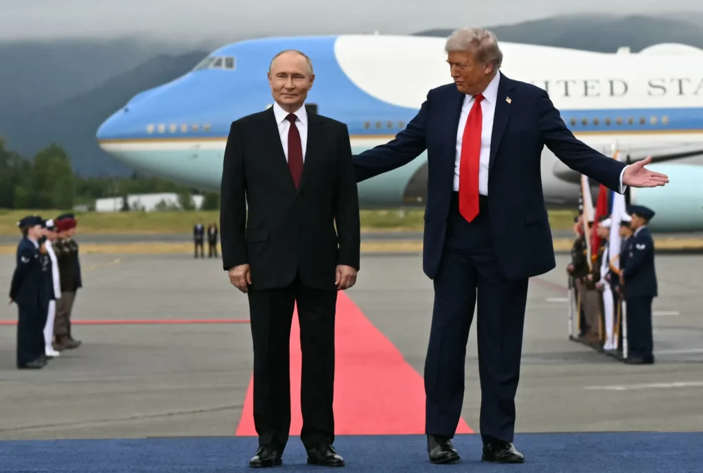 Donald Trump and Vladimir Putin stand on an airport tarmac in front of Air Force One. Trump gestures with an open hand while Putin stands beside him.