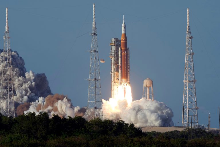 A large orange and white NASA Space Launch System (SLS) rocket lifts off from a launchpad, spewing massive flames and thick white and brown smoke from its base against a clear blue sky. Launch towers and a water tower are visible in the background, with green trees in the foreground.