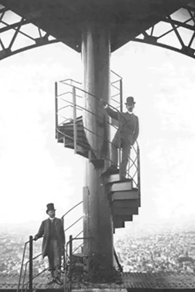 Black and white photo of two men in suits and hats on a spiral staircase, overlooking a city from a high vantage point.
