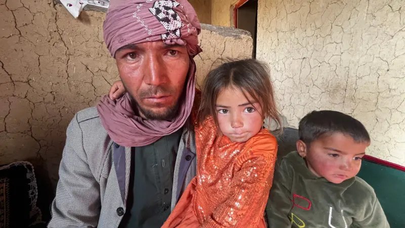 A man in a turban with a young girl in an orange dress and a young boy in a green hoodie, seated against a cracked mud wall.