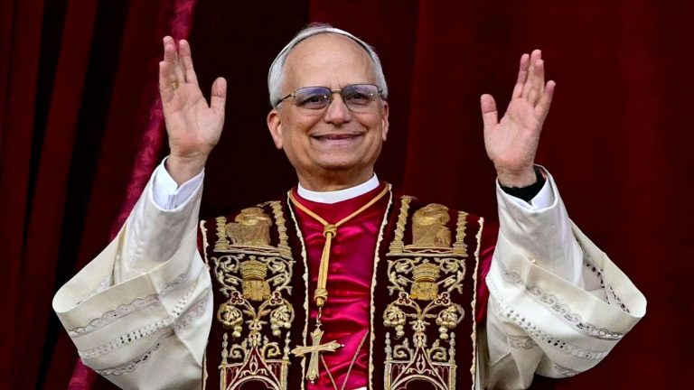 A smiling man in religious vestments and a white skullcap waves with both hands against a red background.